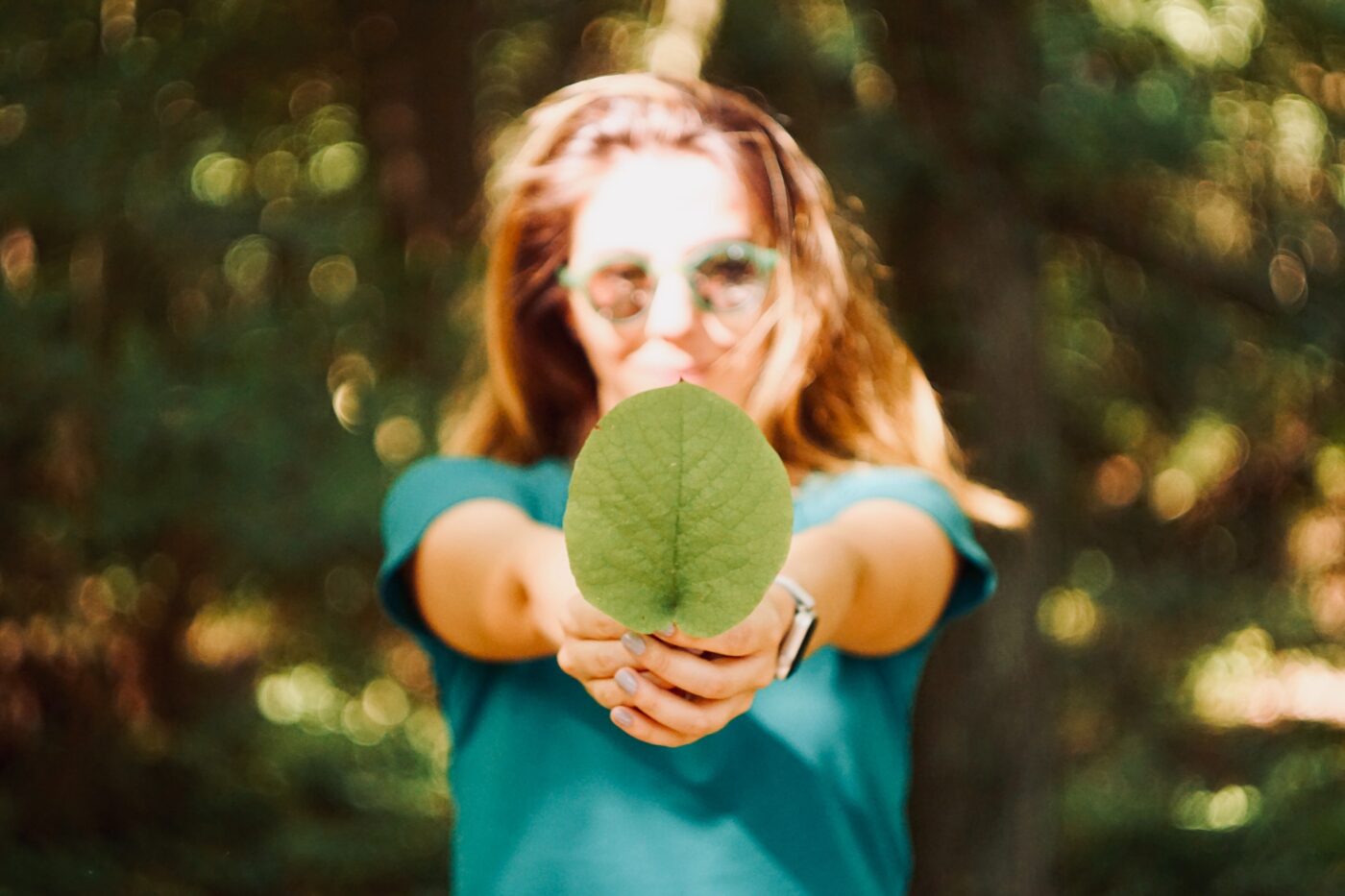 A person wearing a teal shirt and sunglasses holds a large green leaf towards the camera, partially obscuring their face. The background is a lush, sunlit forest.