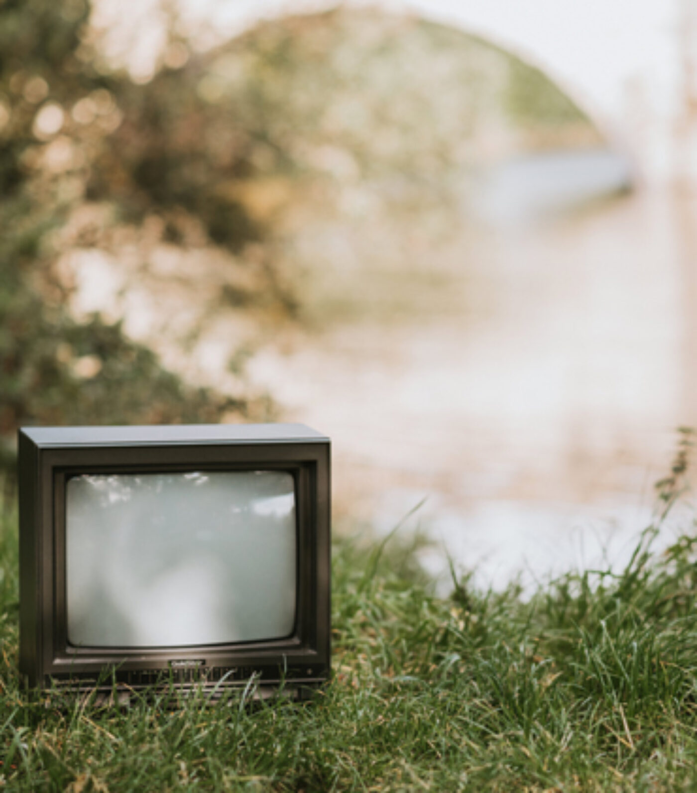 Un vieux petit téléviseur est posé sur l'herbe, près d'un arrière-plan flou et ensoleillé composé d'arbres et d'un pont. Le téléviseur est éteint et son écran est vide, ce qui crée une ambiance nostalgique dans ce décor extérieur.