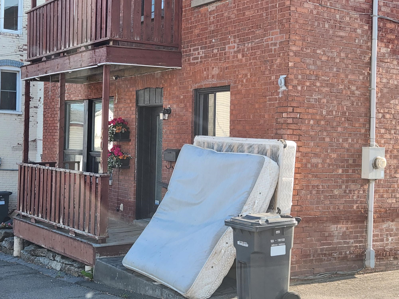 Une maison en briques à deux étages possède un balcon en bois à l'étage supérieur et un porche en dessous. Un matelas et un sommier sont posés contre la rambarde du porche, à côté d'une poubelle extérieure. Des fleurs rouges sont suspendues dans des paniers près de la porte d'entrée.