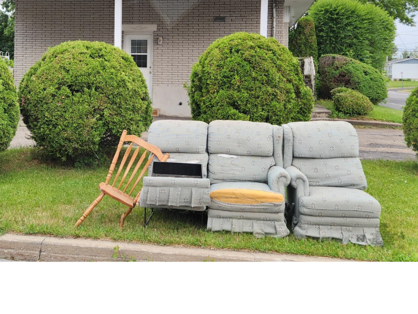 Bulky items on a lawn in front of a house with bushes in the background. The items are a grey worn armchair and a broken chair.