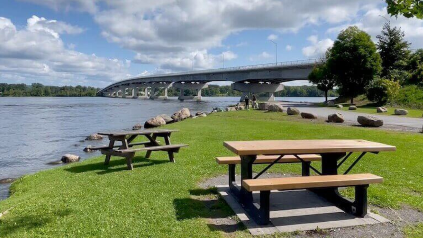 Picnic tables on a grassy riverbank with a bridge in the background under a partly cloudy sky.